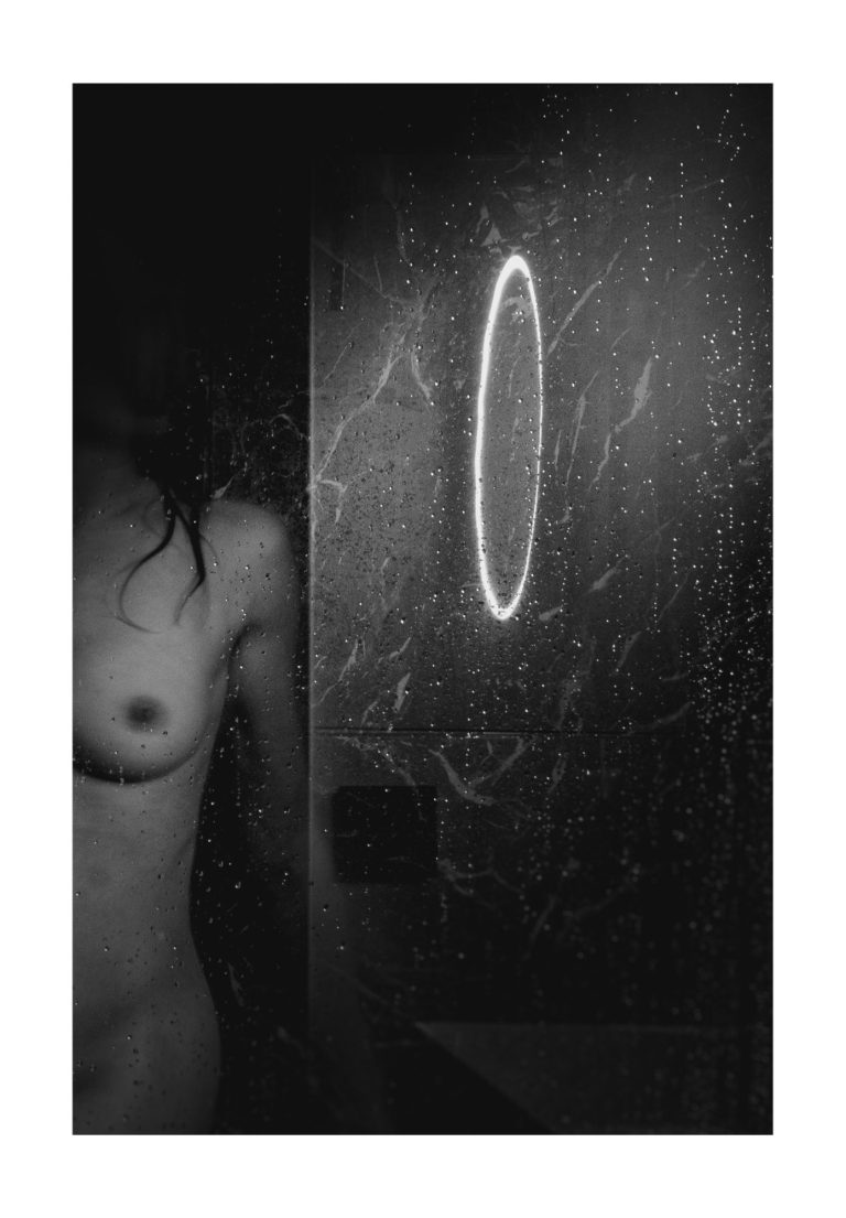 Naakt figuur gedeeltelijk zichtbaar naast een beslagen spiegel met een ronde reflectie.