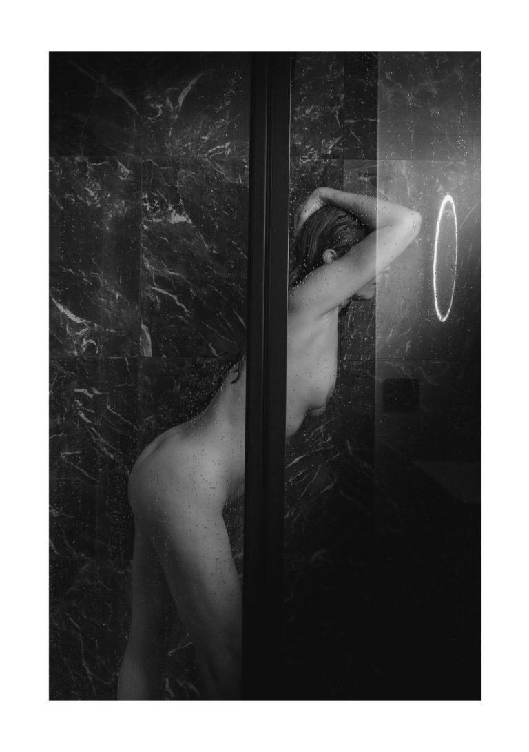 Naakte vrouw staat in een douche, gedeeltelijk zichtbaar achter een glazen wand.